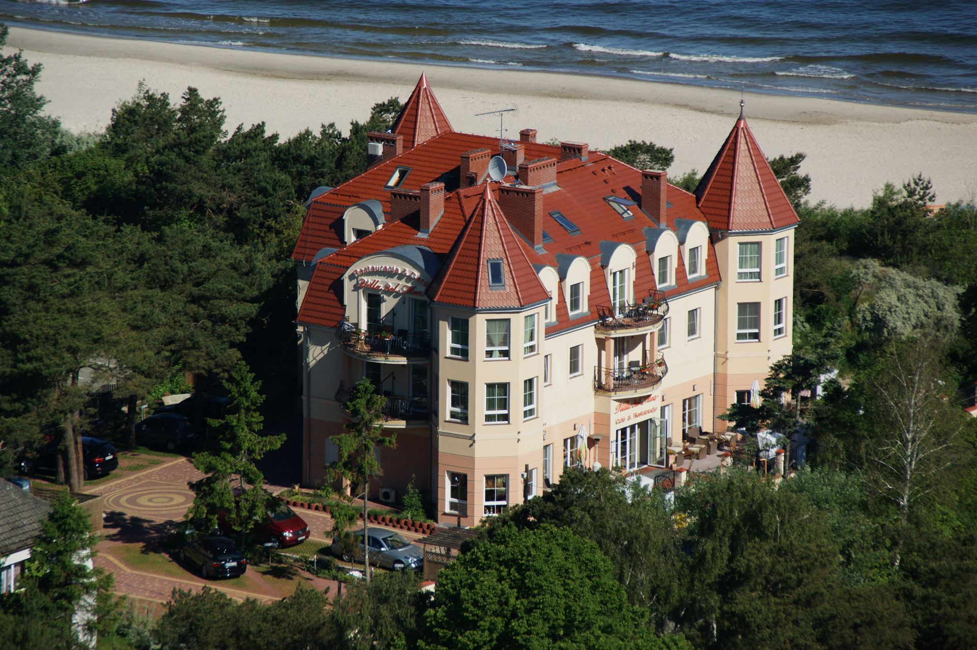Budynek hotelu z czerwonym dachem i wieżyczkami pośród drzew, przy piaszczystej plaży