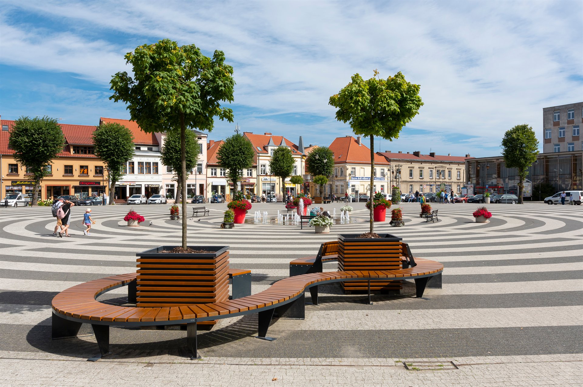 Miejski rynek z okrągłym placem, drzewami w donicach i ławkami, otoczony kamienicami z czerwonymi dachami
