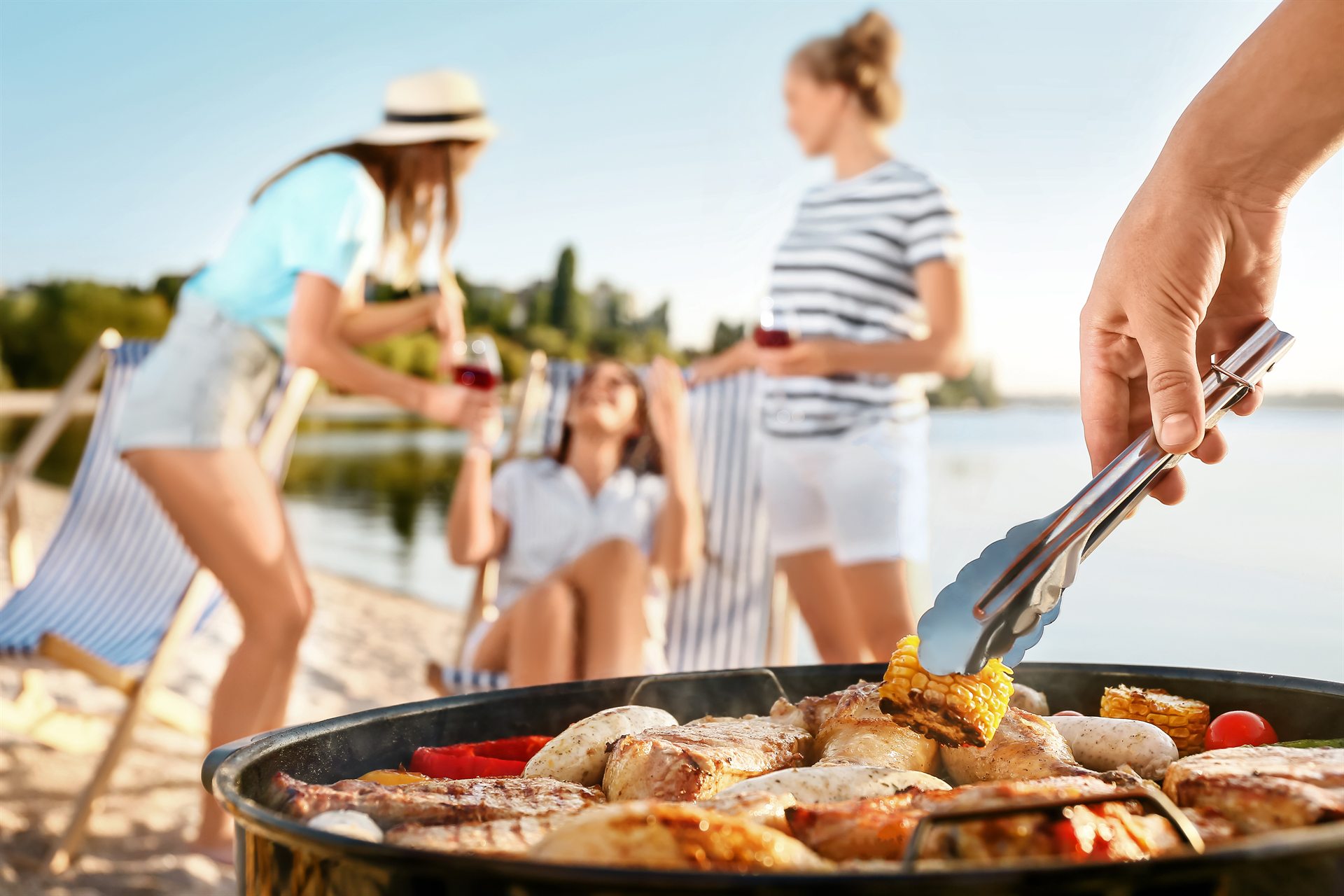 Letnie grillowanie nad wodą, w tle rozmyte osoby z kieliszkami, na pierwszym planie ręka obraca kukurydzę szczypcami