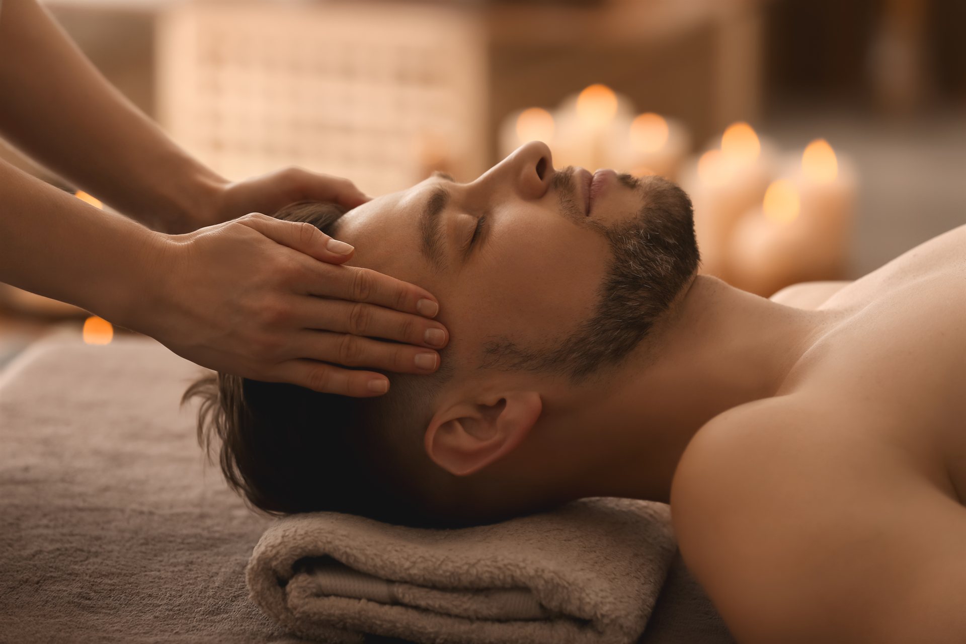 Mężczyzna leży na stole do masażu, a terapeuta wykonuje relaksujący masaż głowy wśród świec