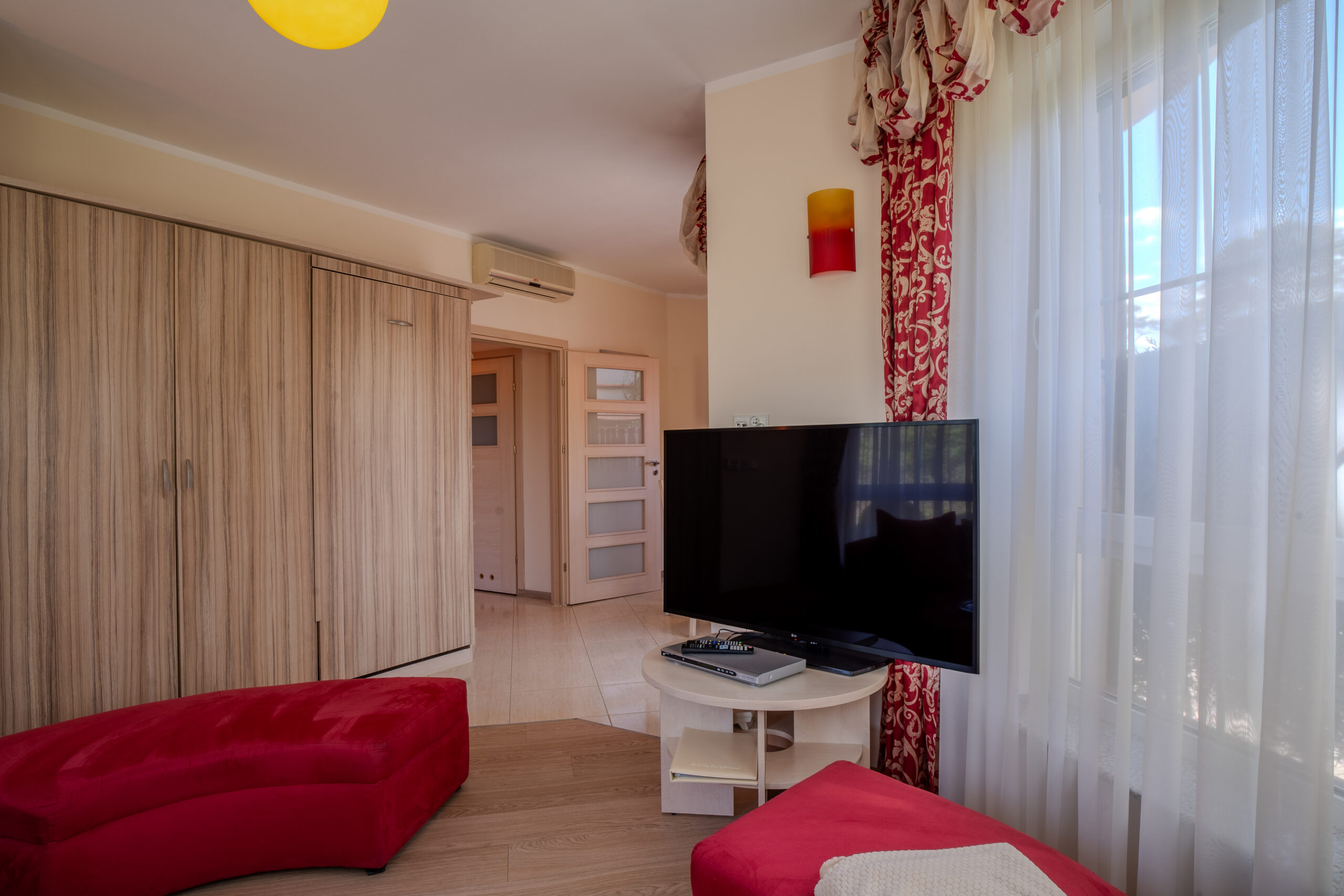 Nowoczesny salon z czerwonymi pufami, telewizorem przy oknie i dużą szafą na jasnym tle