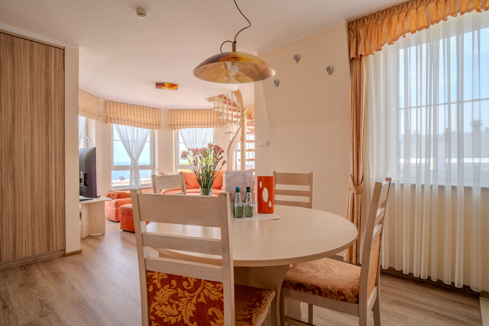 Jasny salon z jadalnią, okrągłym stołem, pomarańczową sofą i spiralnymi schodami przy oknie