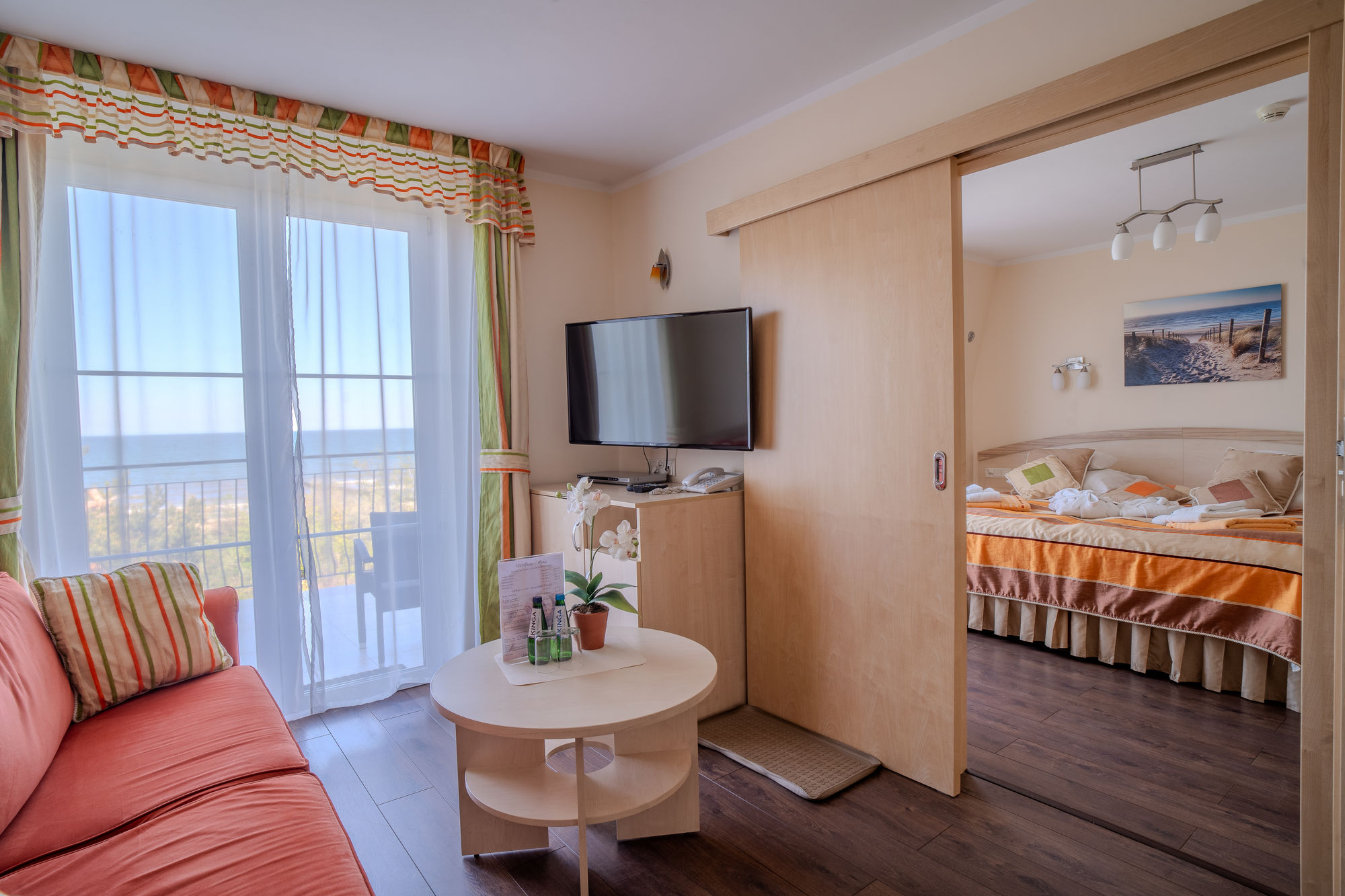 Jasny pokój hotelowy z sofą, telewizorem, łóżkiem i widokiem na morze przez balkon