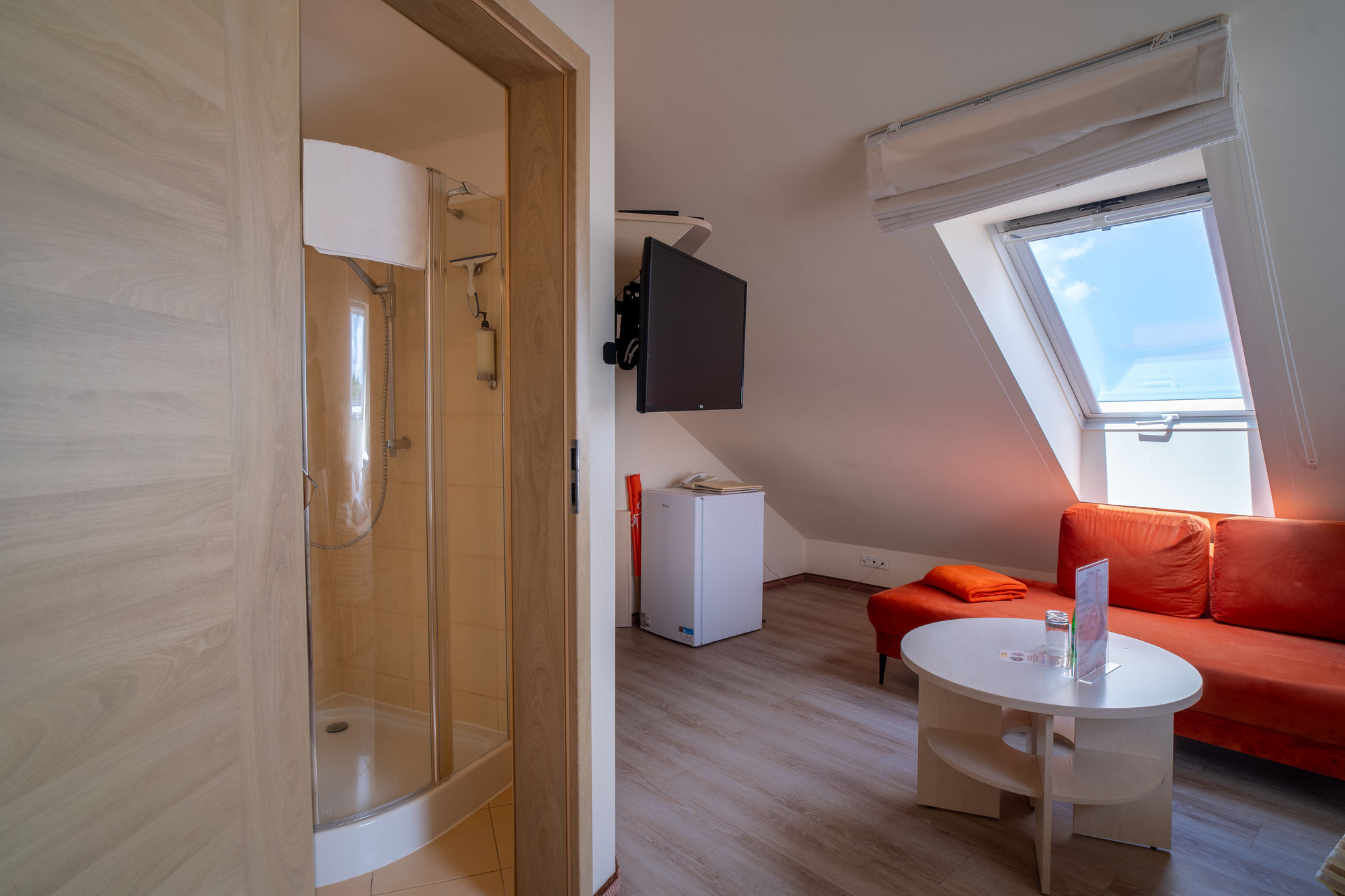 Jasny pokój hotelowy z łazienką, pomarańczową sofą, TV i oknem dachowym