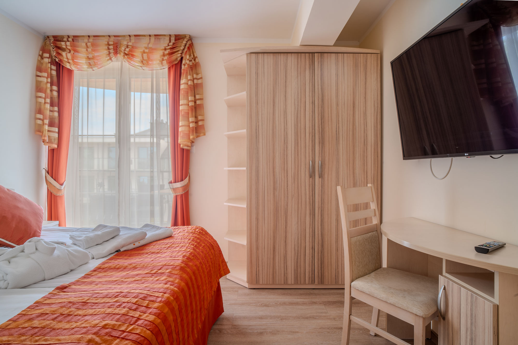 Jasny pokój hotelowy z łóżkiem, szafą, biurkiem, telewizorem i oknem z pomarańczowymi zasłonami