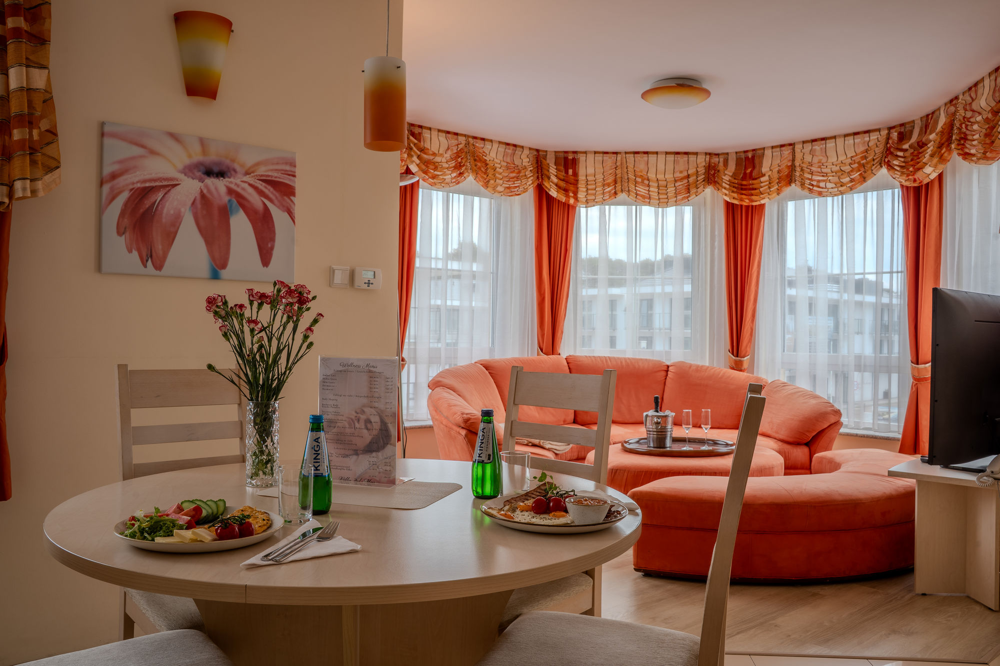 Przytulny salon z pomarańczową sofą, nakrytym stołem, kwiatami i dużymi oknami z zasłonami