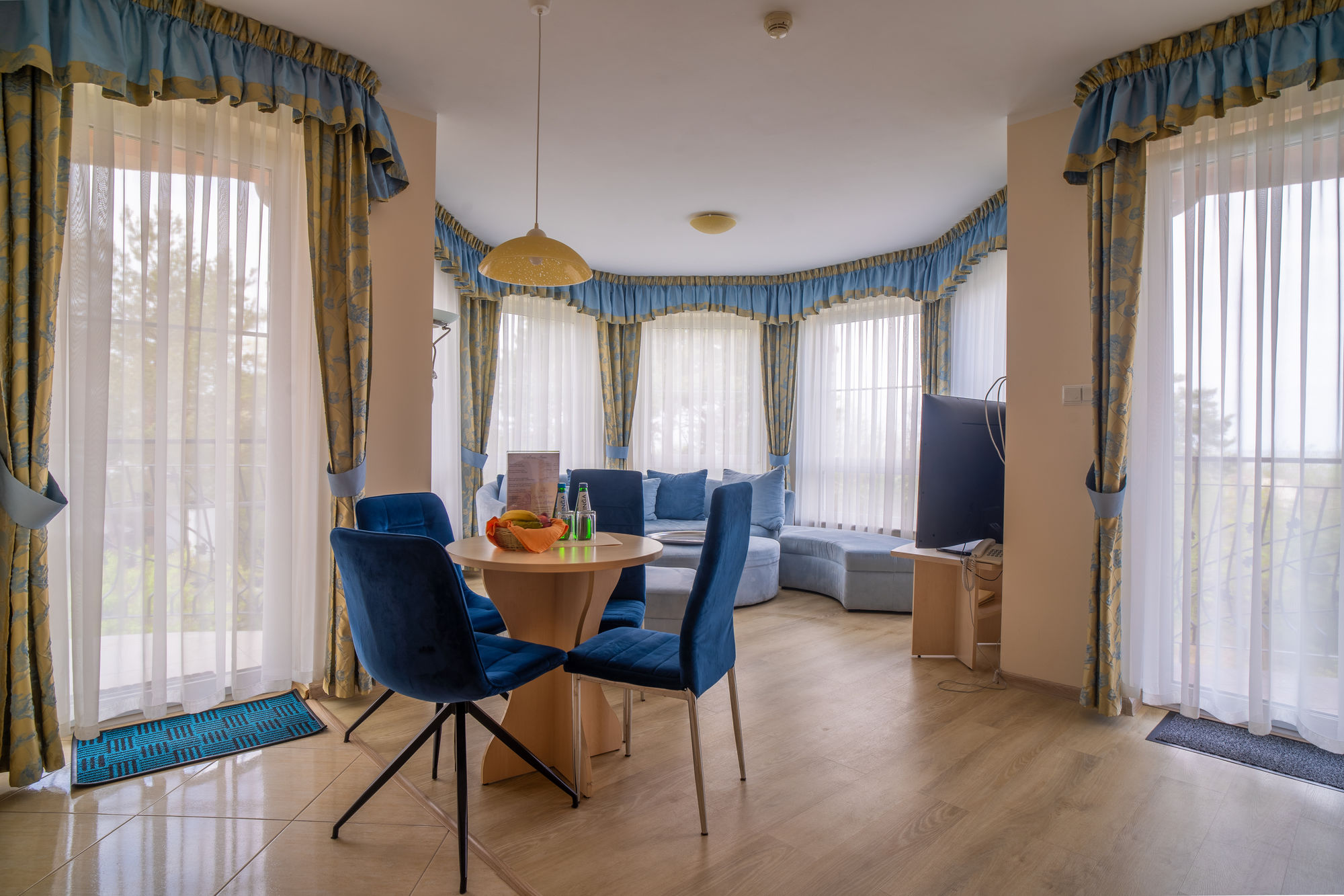 Jasny salon z niebieską sofą, stołem i krzesłami, otoczony dużymi oknami z firanami i zasłonami
