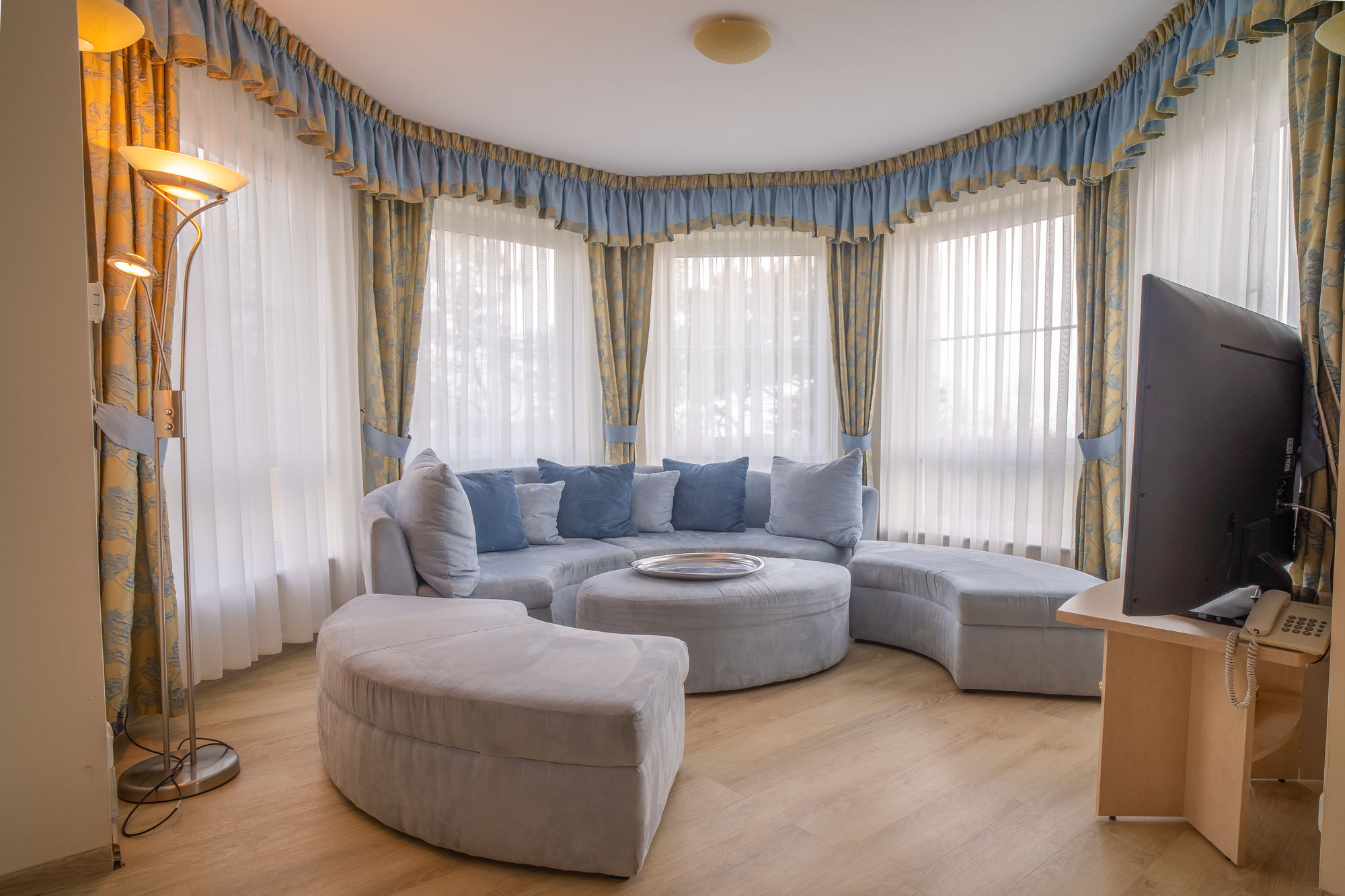 Jasny salon z narożną kanapą, pufą, lampą stojącą i telewizorem przy dużych oknach