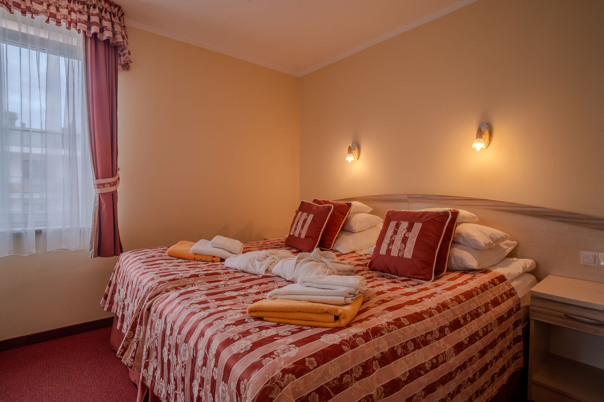 Przytulny hotelowy pokój z podwójnym łóżkiem, czerwonym narzutą, ręcznikami i ciepłym oświetleniem