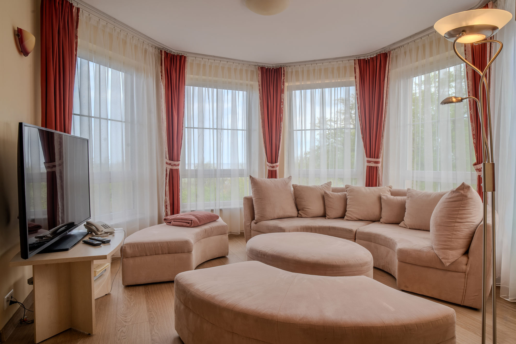 Jasny salon z beżową narożną sofą, dużymi oknami z czerwonymi zasłonami i telewizorem