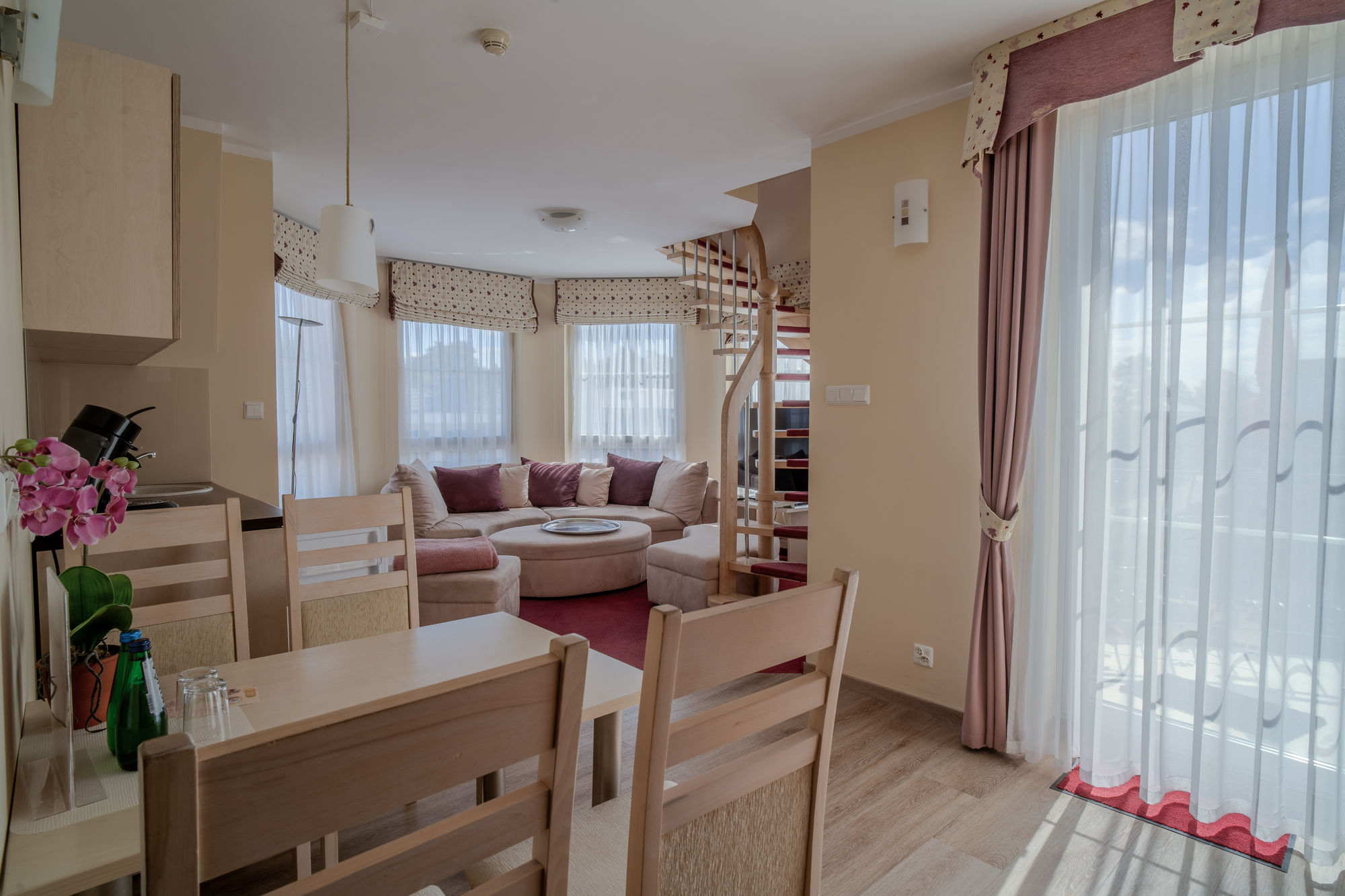 Jasny salon z jadalnią, beżowa sofa, okrągłe okno i spiralne schody przy dużych oknach