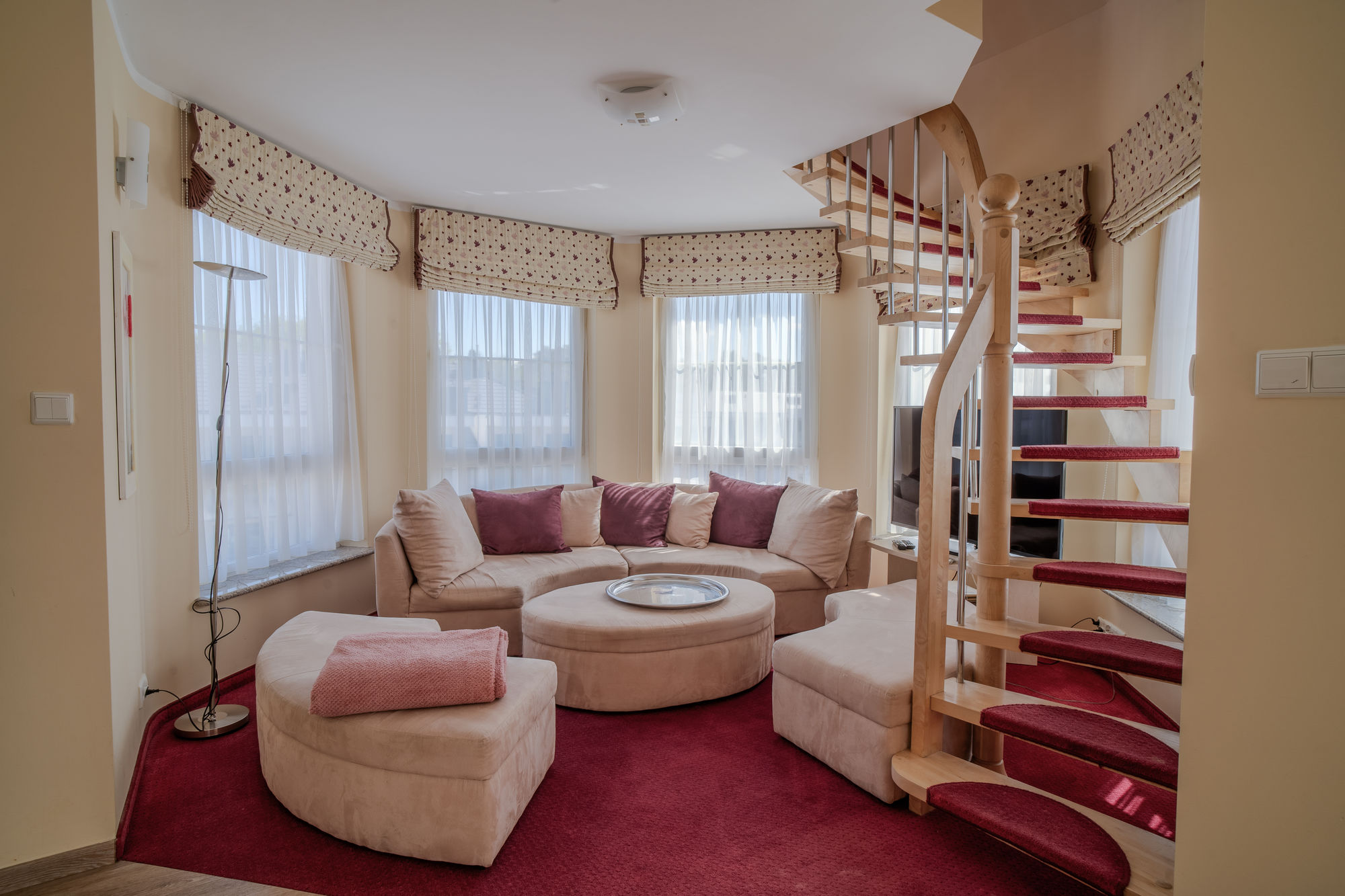 Przytulny salon z beżową sofą, bordowym dywanem i spiralnymi schodami z czerwonymi stopniami