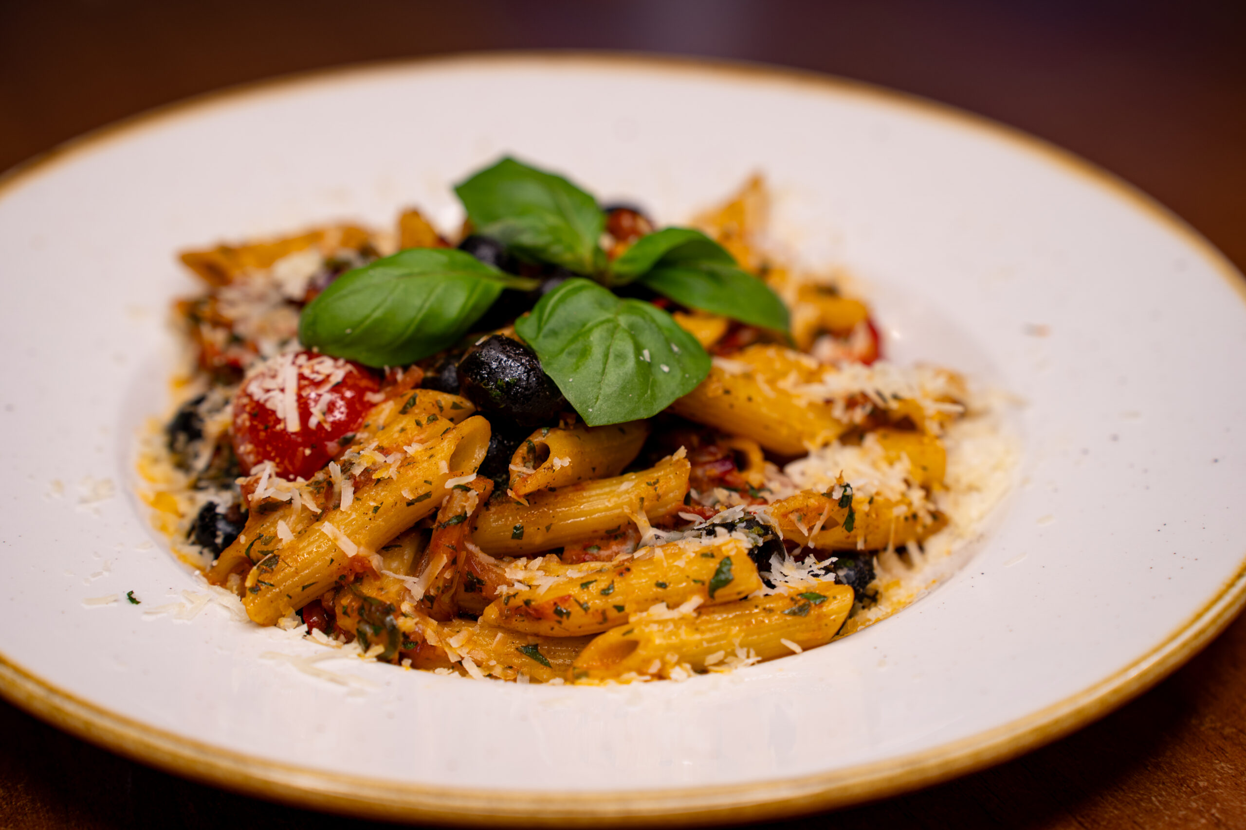 Makaron penne z pomidorami, czarnymi oliwkami, parmezanem i świeżą bazylią na białym talerzu