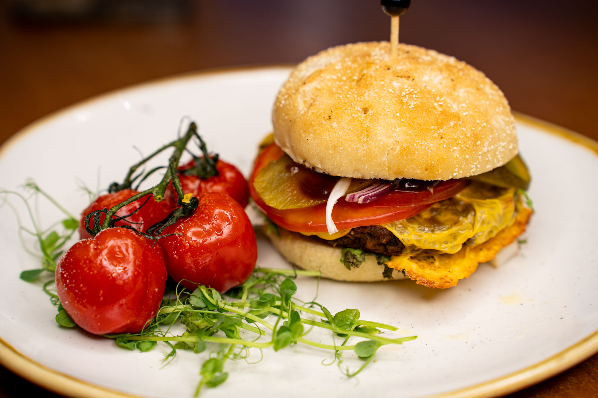 Burger z warzywami na białym talerzu, obok gałązka czerwonych pomidorów i zielone kiełki